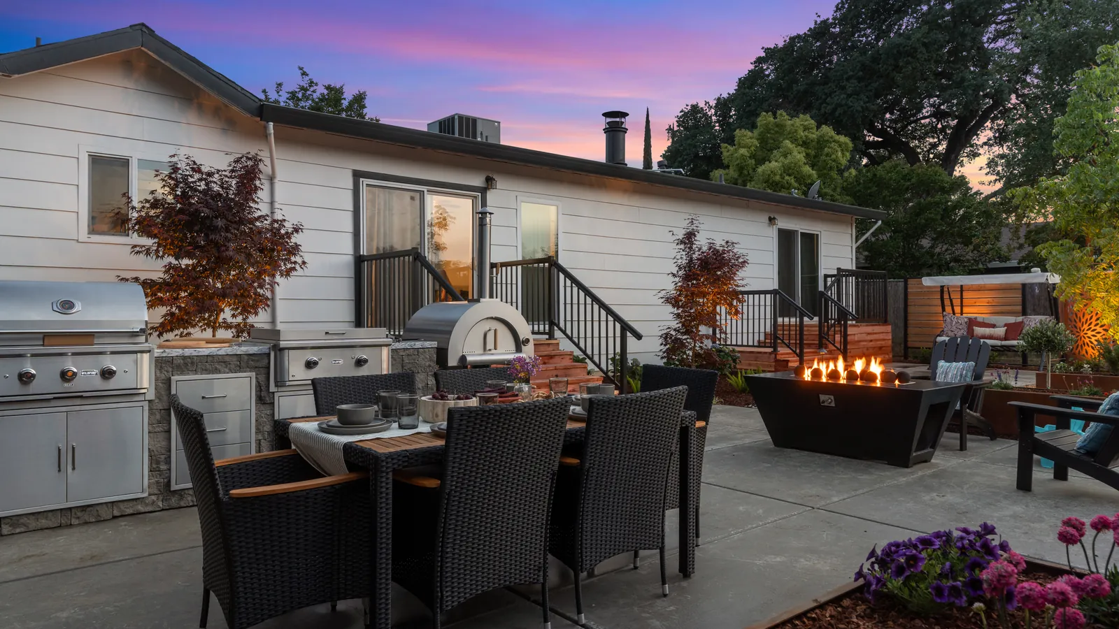 Outdoor kitchen, dining patio, and fire table at dusk in a backyard by Stuteville Landscaping
