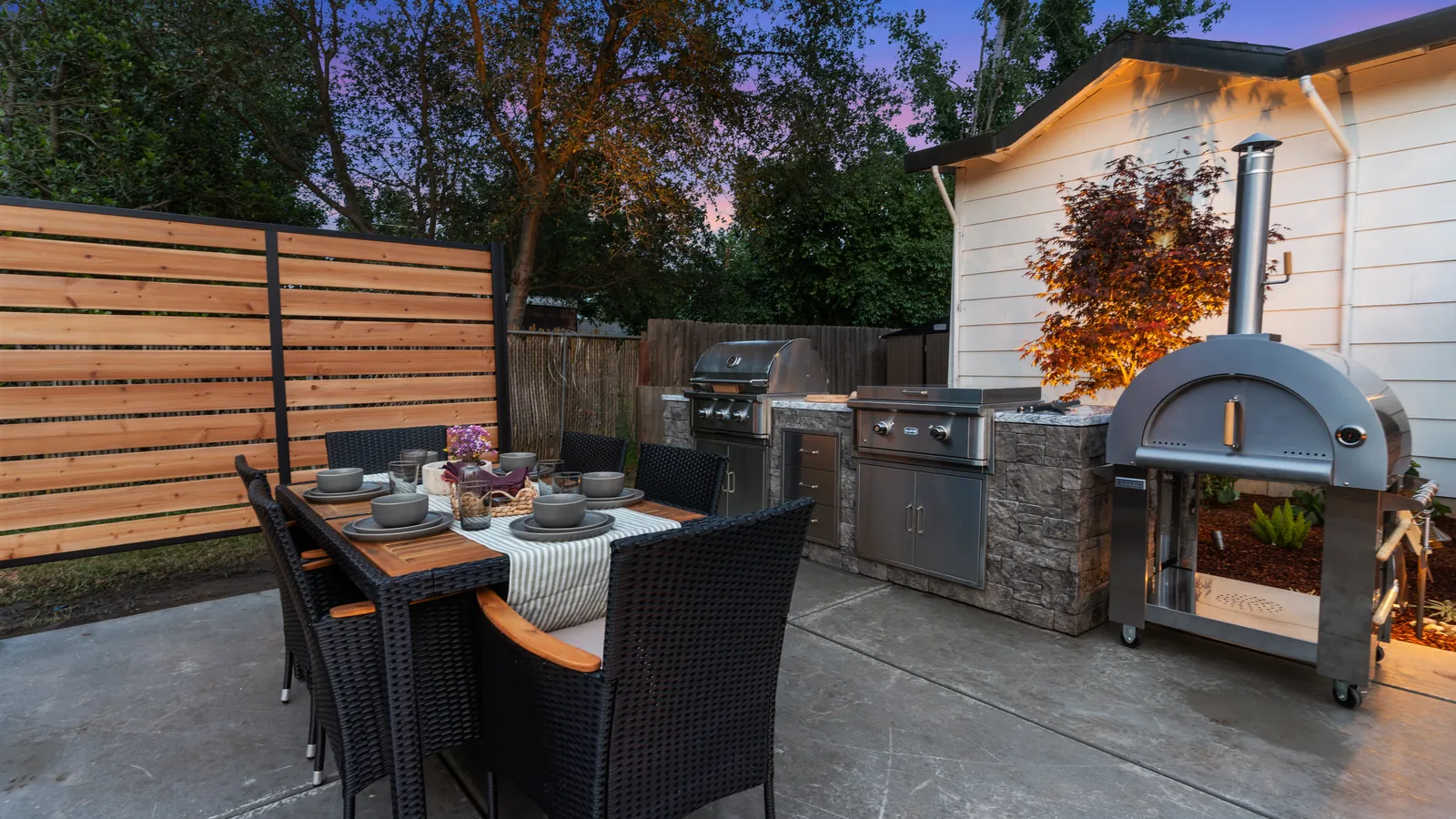 Outdoor kitchen with built-in grills and a pizza oven beside a dining table at dusk
