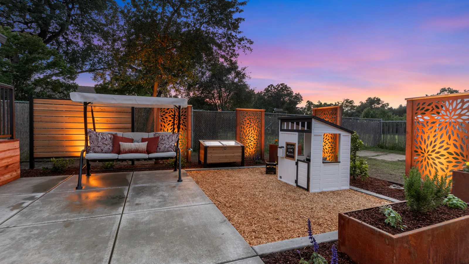 Covered swing, playhouse, and corten planters on pea gravel at dusk